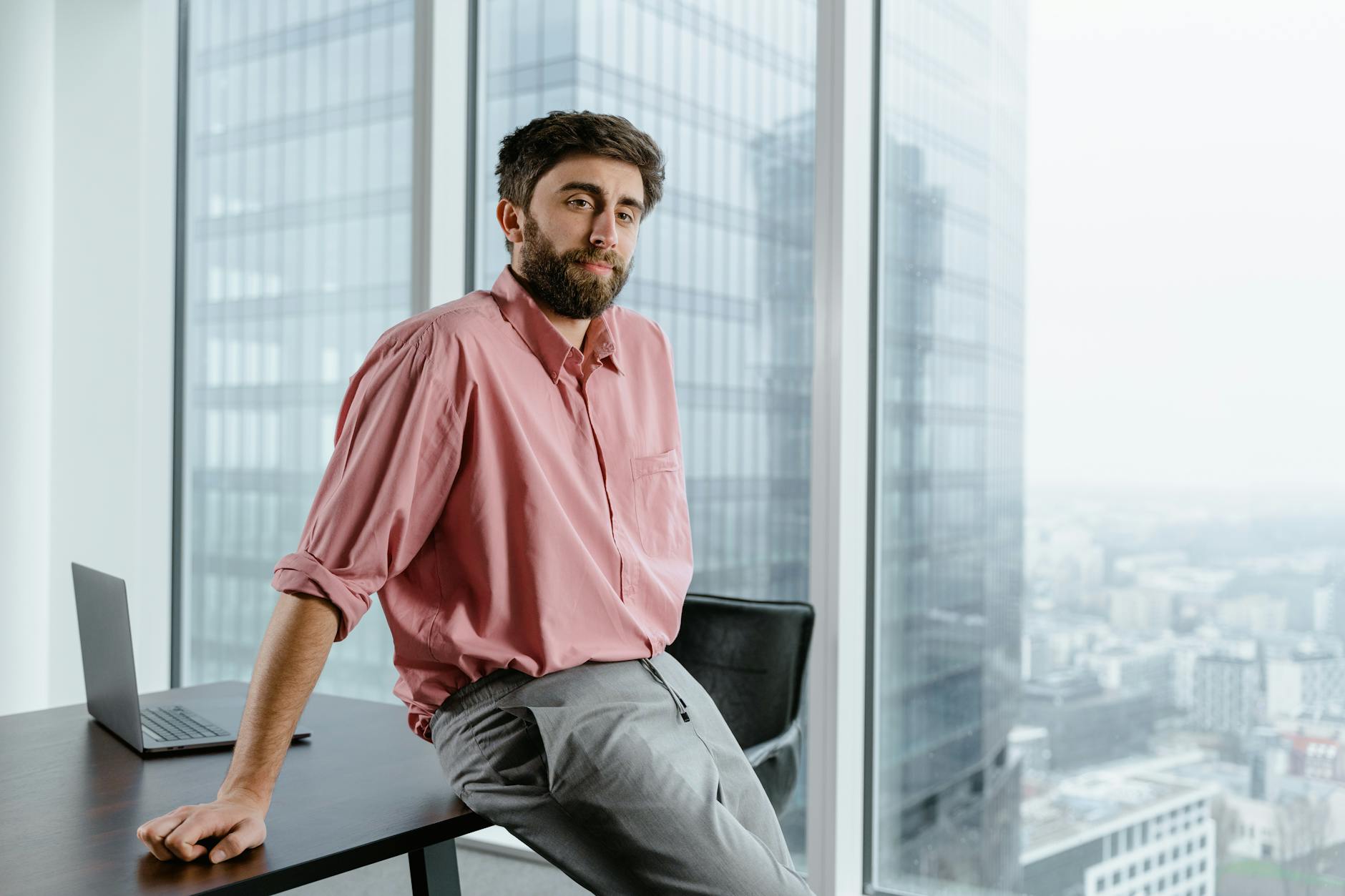 A businessman with facial hair in a pink shirt sits by a large window in a modern office building.