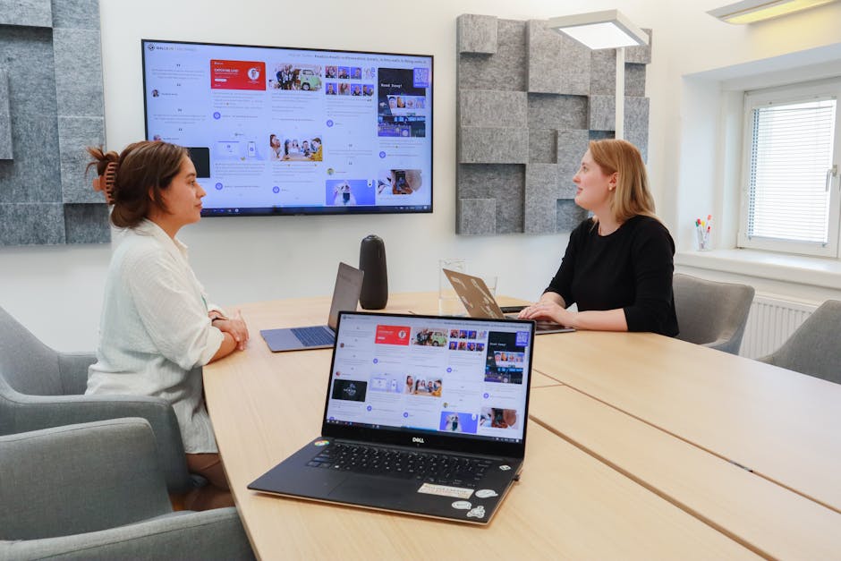 Women collaborating in a modern office with laptops and large digital display.